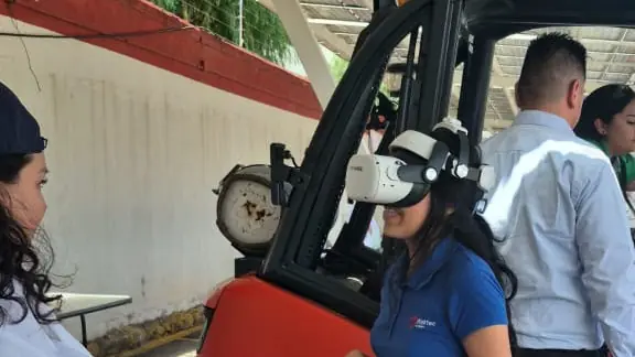 Participante con visor de realidad virtual junto a un montacargas rojo bajo un techo de paneles solares, mientras otras personas observan