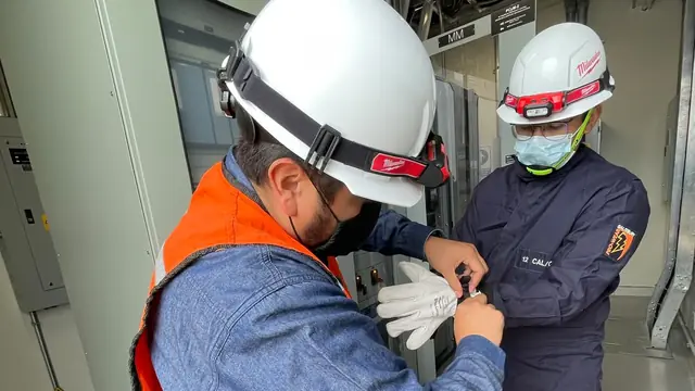 Técnico con casco blanco y chaleco naranja ajusta el equipo de un compañero junto a tableros eléctricos en un cuarto de máquinas