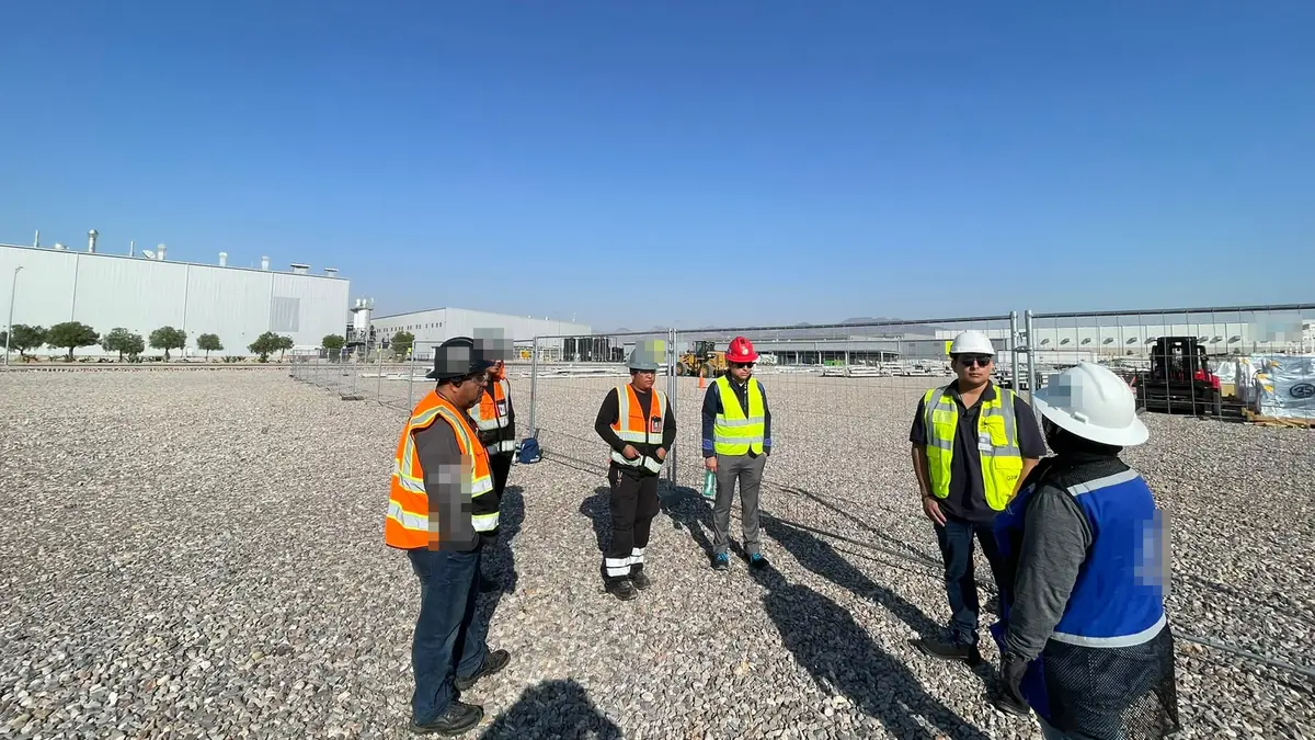 Seis personas con casco y chaleco de alta visibilidad reunidas en un patio de grava frente a las naves de una planta industrial