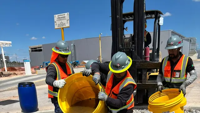 Tres trabajadores con casco y chaleco vacían cubetas de líquido en un registro subterráneo abierto, con un montacargas detrás