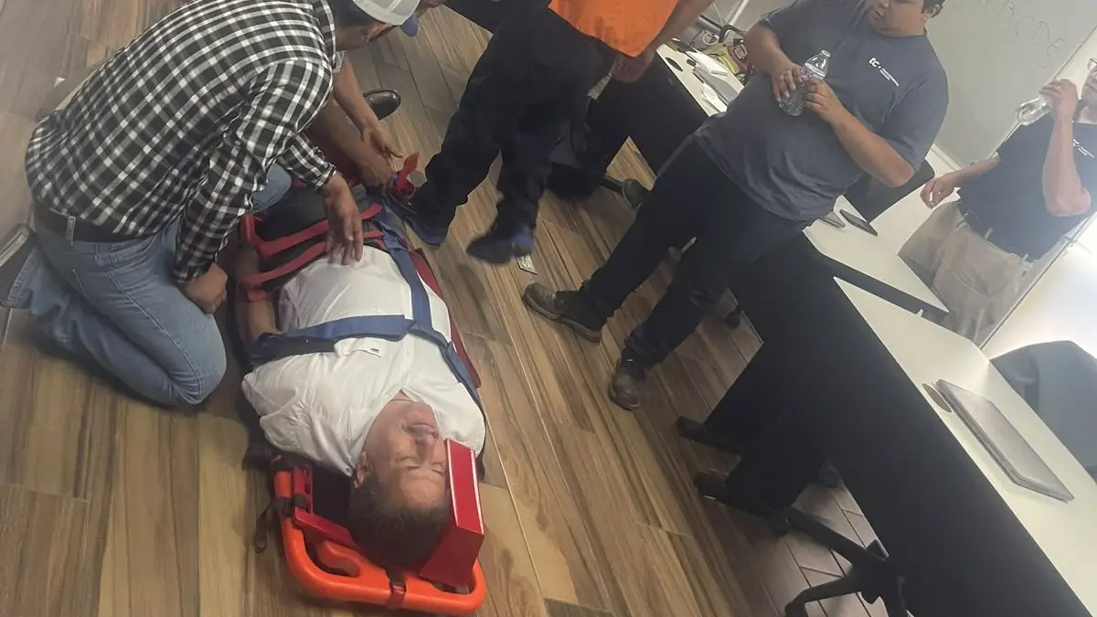 Grupo en un aula practicando primeros auxilios con un participante inmovilizado en una camilla naranja