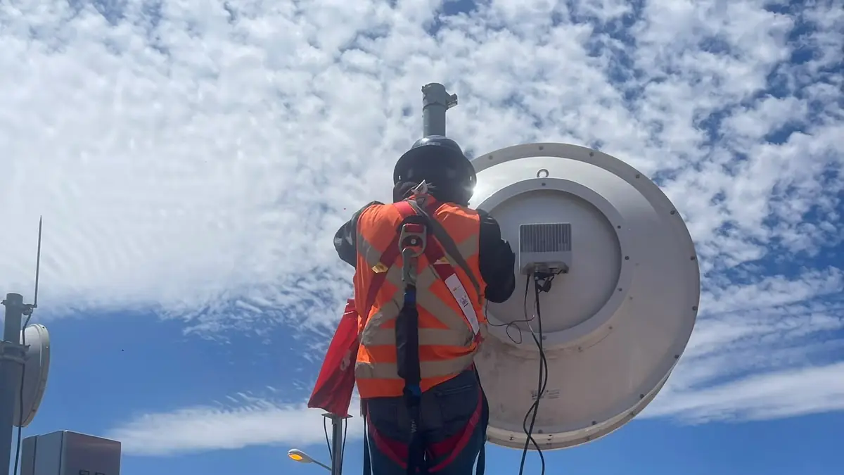 Técnico con arnés de cuerpo completo y chaleco naranja de pie sobre un mástil en la azotea de un edificio, junto a una antena y un tablero eléctrico
