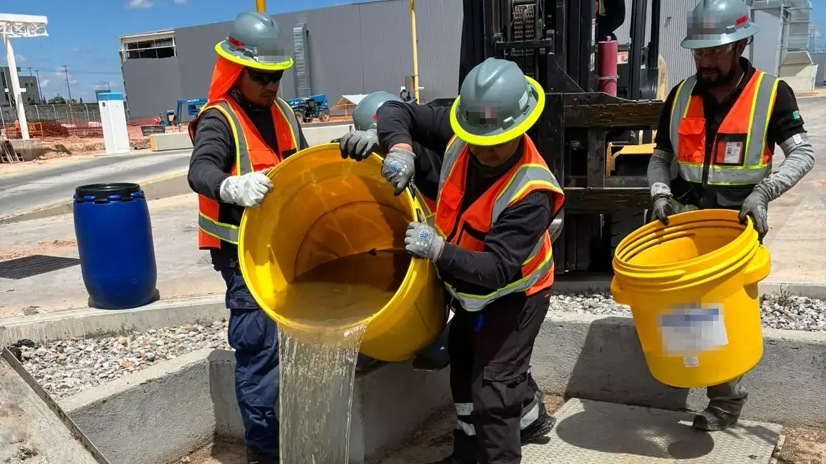 Three workers in hard hats, high-visibility vests and gloves handle yellow buckets beside an open utility vault, two of them tipping one bucket together