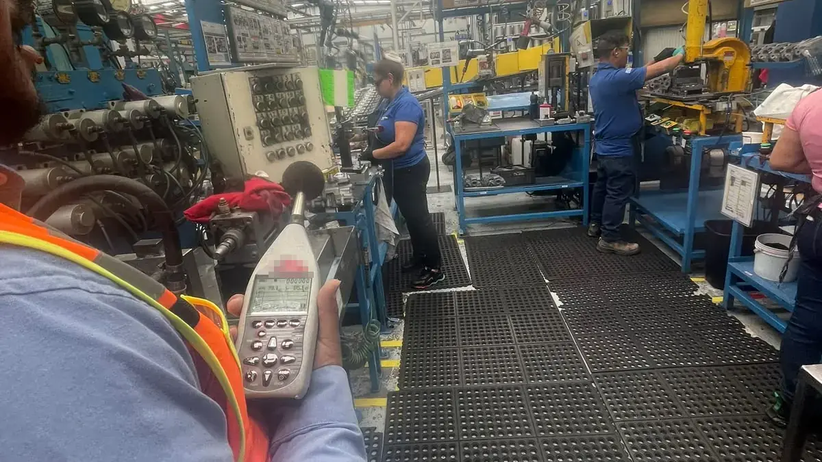 Technician in an orange vest holding a sound level meter beside a production line while two operators work at their stations on anti-fatigue mats
