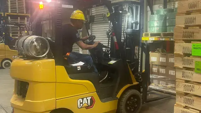 Operator in a hard hat driving a Cat forklift down a warehouse aisle lined with racks