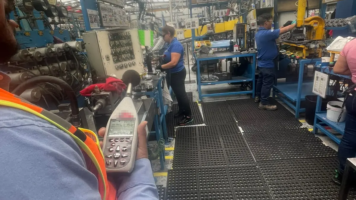 Técnico con chaleco naranja sostiene un sonómetro junto a una línea de producción mientras dos operadores trabajan en sus estaciones sobre tapetes antifatiga