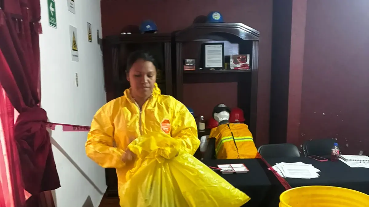 Participante con traje amarillo de protección química sostiene una bolsa del kit de derrames junto a una cubeta, en un aula acordonada