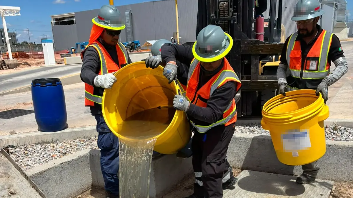 Tres trabajadores con casco, chaleco reflejante y guantes manejan cubetas amarillas junto a un registro abierto: dos vacían una entre ambos y el tercero sostiene otra