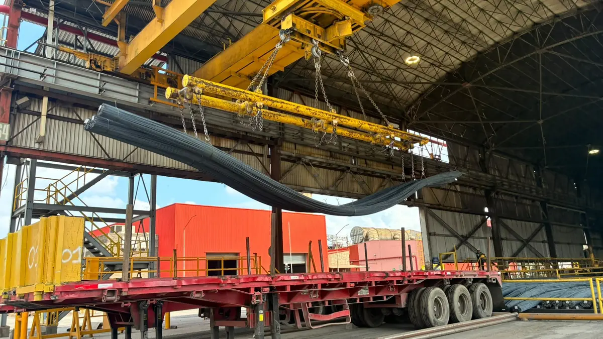 Grúa viajera baja un atado de varillas de acero sobre un tráiler en el andén de una nave industrial