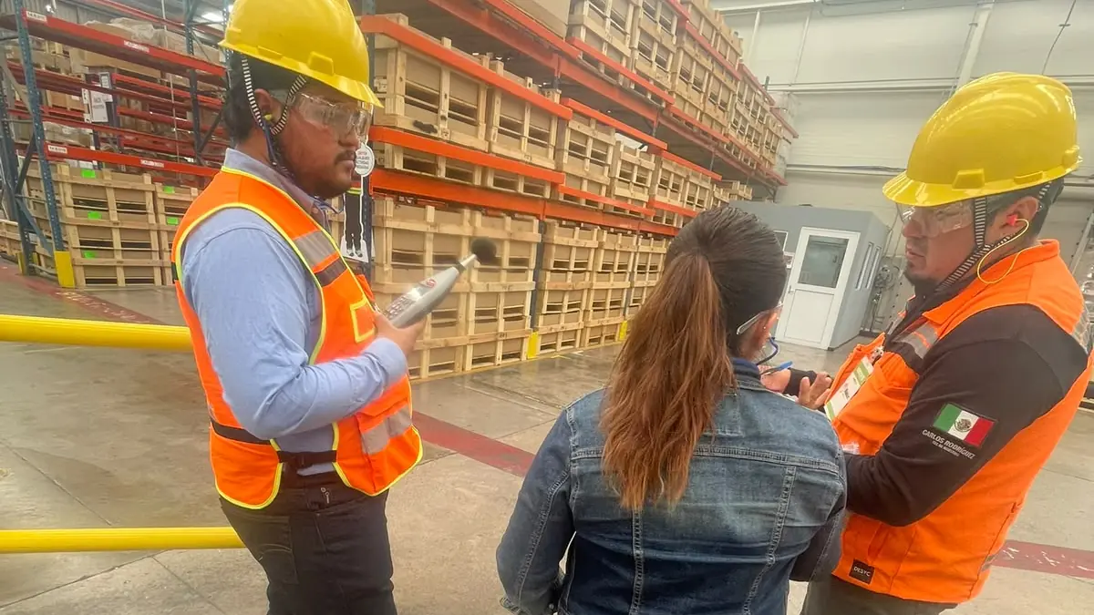 Supervisor de seguridad con chaleco dando indicaciones a dos trabajadores en el piso de planta