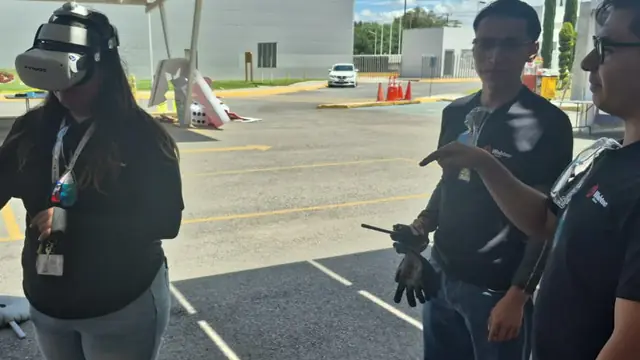 Participante con visor de realidad virtual en una demostración del simulador de montacargas, al aire libre bajo un techo de paneles solares, mientras dos instructores con playera negra observan y uno señala