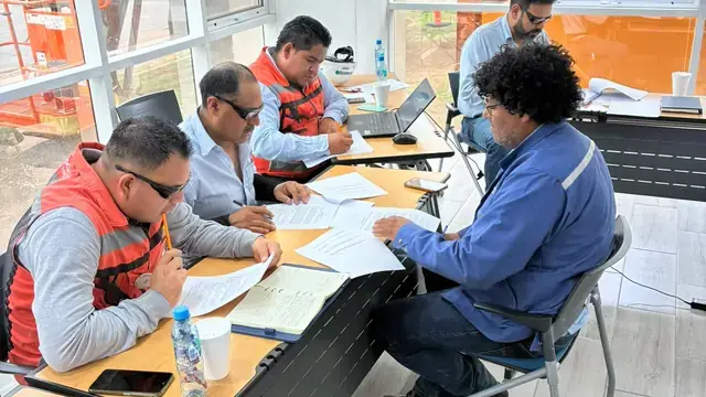 Two supervisors in safety vests go through binders and forms at a desk in a plant office