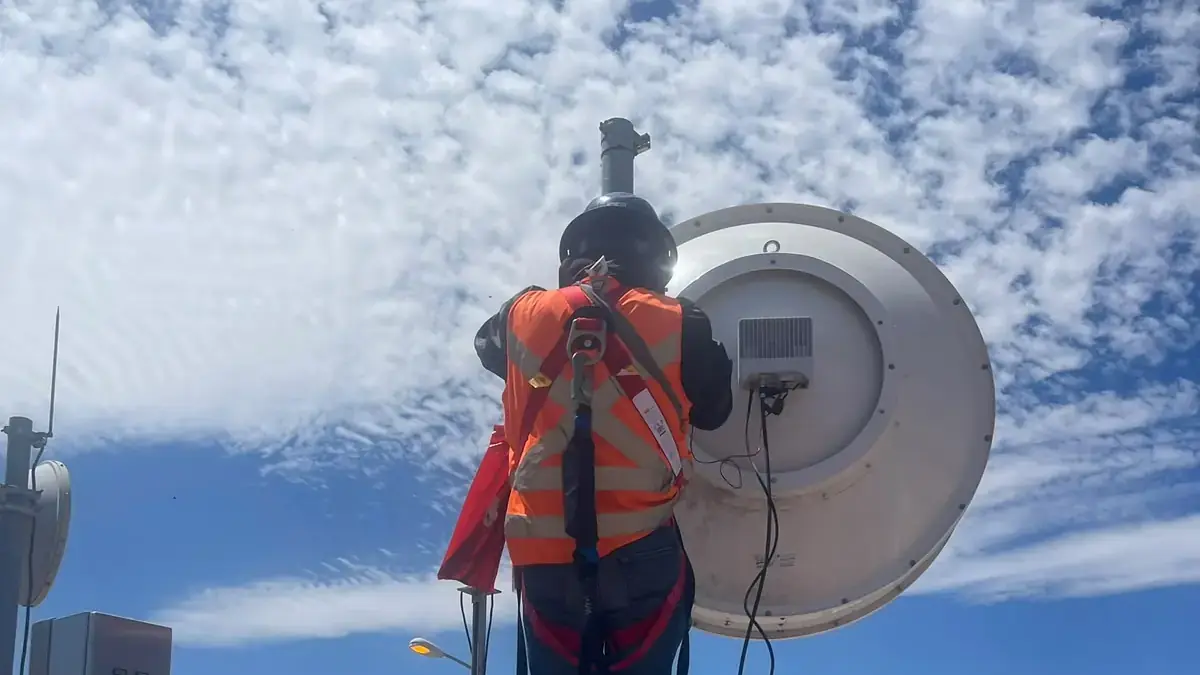 Technician in a full-body harness and orange vest standing on a rooftop mast next to an antenna and an electrical panel