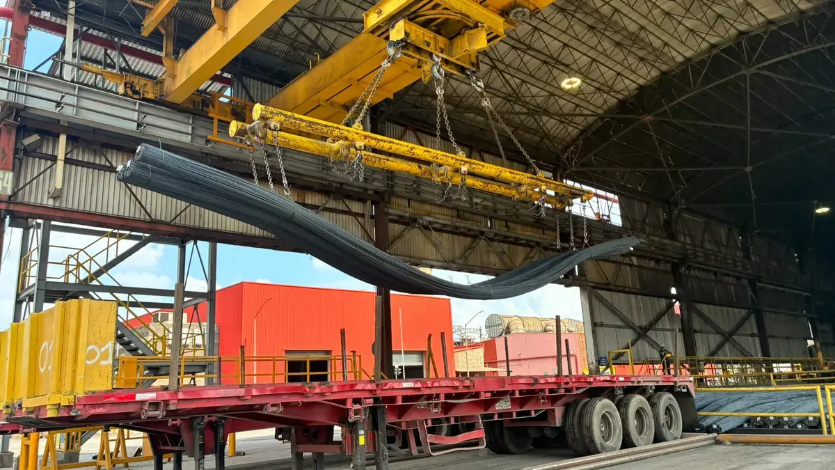 Overhead crane lowering a bundle of steel bars onto a flatbed trailer at the edge of an industrial building
