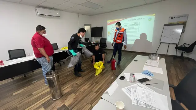 Aula de capacitación con participantes de pie, un extintor junto a las mesas y la presentación del curso proyectada al fondo