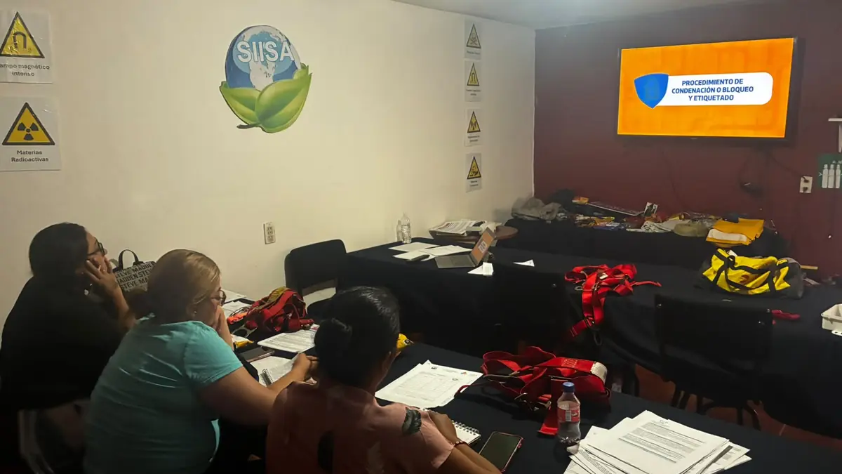 Participantes en el aula de SIISA con arneses y hojas impresas sobre las mesas, frente a una pantalla con un procedimiento de bloqueo y etiquetado