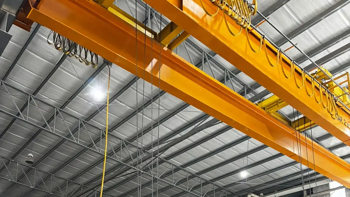 Factory bay with light fixtures hanging from a high roof and a worker in a hard hat checking tags on stacked steel bars under an overhead crane