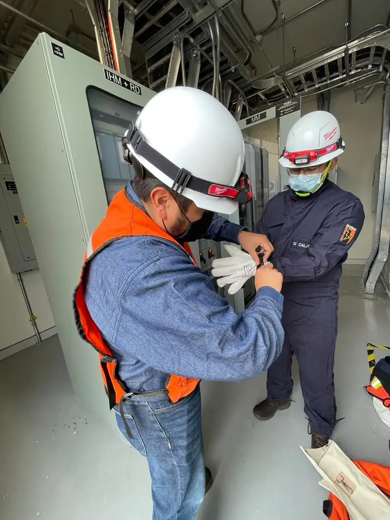 Técnico con casco blanco y chaleco naranja ajusta el equipo de un compañero junto a tableros eléctricos en un cuarto de máquinas