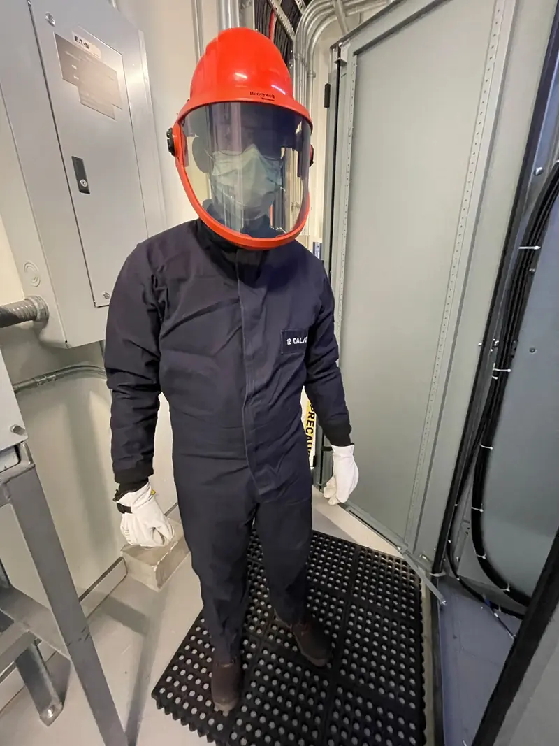 Técnico con traje de protección contra arco eléctrico, careta y guantes, de pie entre tableros eléctricos