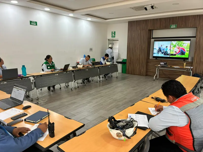 Sala de capacitación dentro de una planta: participantes con chaleco y casco toman notas frente a la pantalla del curso
