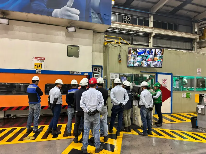 Trabajadores con casco y chaleco en una sesión de seguridad junto a una línea de prensas