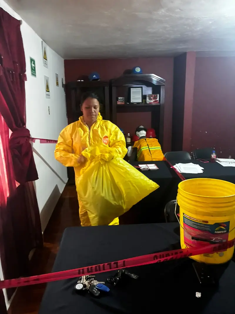 Participante con traje amarillo de protección química sostiene una bolsa del kit de derrames junto a una cubeta, en un aula acordonada