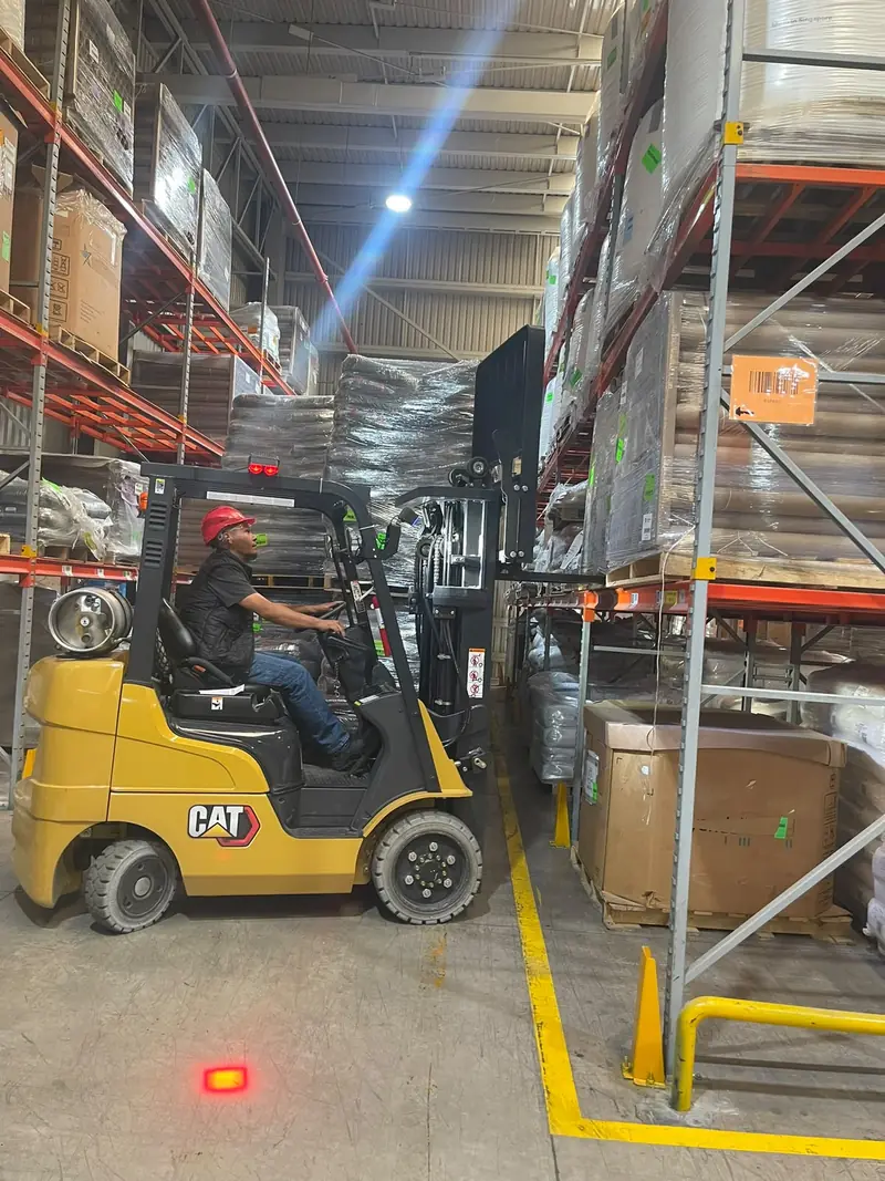 Operador con casco y chaleco conduce un montacargas Cat entre racks de almacén durante la práctica