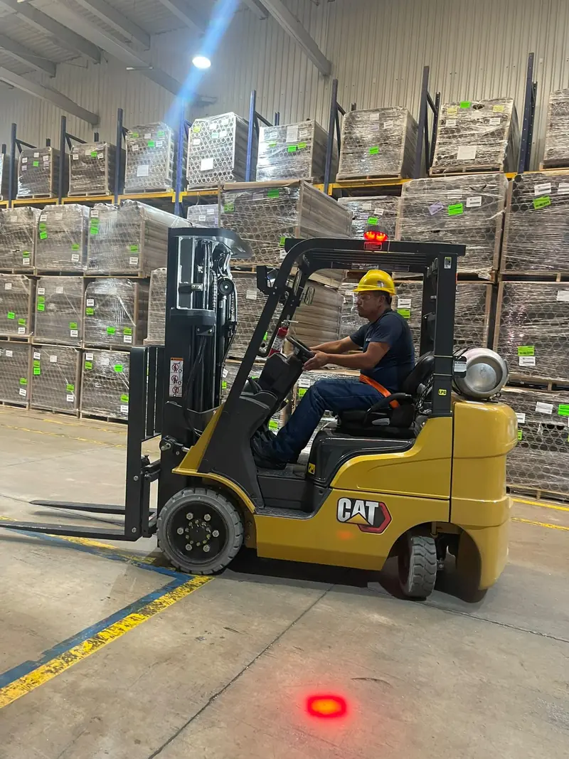 Operador con casco amarillo conduce un montacargas Cat entre racks de almacén durante una práctica