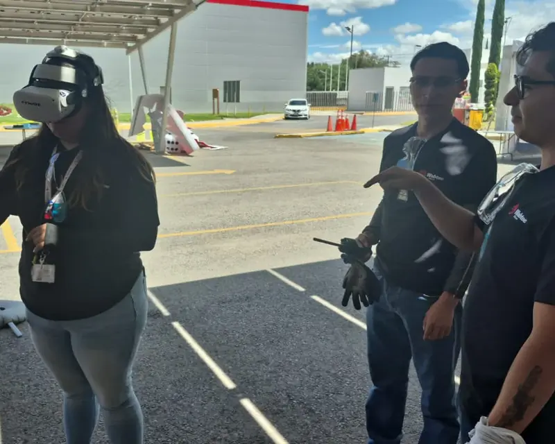 Participante con visor de realidad virtual en una demostración del simulador de montacargas, mientras dos instructores observan y uno le indica la maniobra
