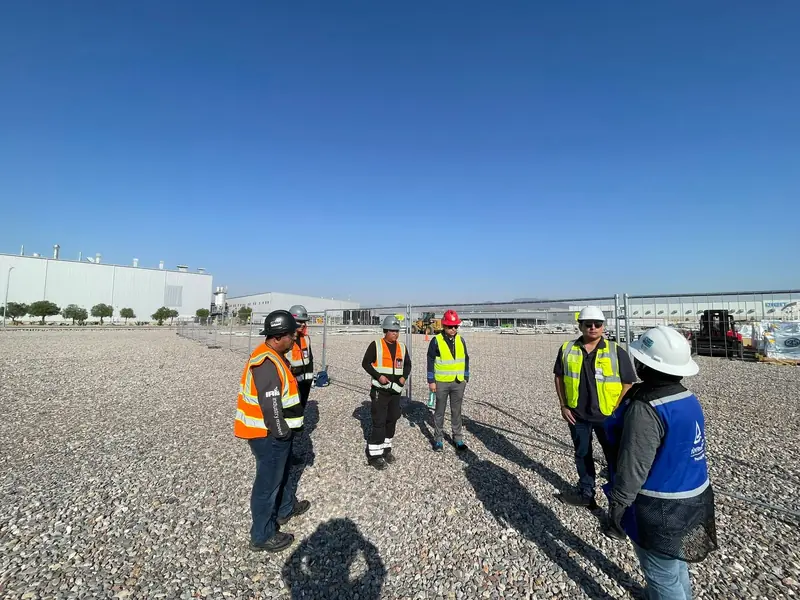 Equipo con chalecos y cascos en un recorrido de seguridad sobre el terreno de la obra, con naves industriales al fondo